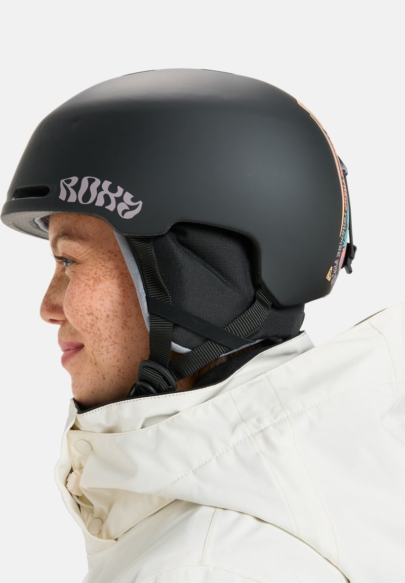 Čierna lyžiarska prilba s nastaviteľnými popruhmi, vnútorným polstrovaním a logom na strane. Nositeľ má na sebe bielu bundu s kapucňou.