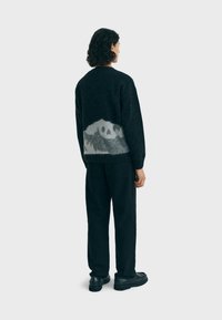 Maglione nero con un design montano testurizzato grigio sul retro. Il modello lo abbina a pantaloni e scarpe nere.