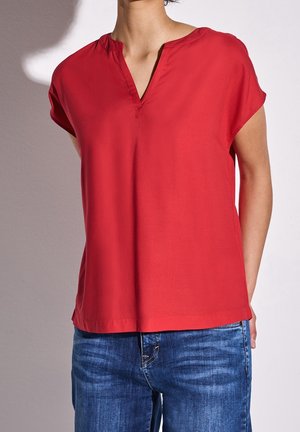 Femme portant un chemisier rouge ample à manches courtes avec un décolleté en V, associé à un jean bleu délavé de longueur moyenne, sur un fond uni.