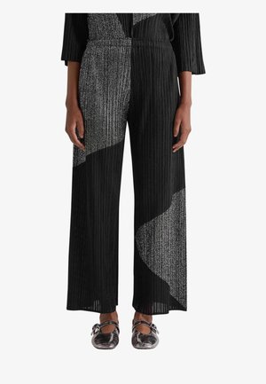 Donna che indossa pantaloni neri plissettati a gamba larga con pannelli diagonali argentati scintillanti, abbinati a scarpe Mary Jane color argento metallizzato.