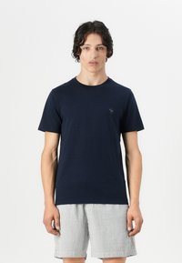 T-shirt in cotone blu navy con colletto rotondo e maniche corte, con un piccolo logo sul petto. Il modello indossa pantaloni corti a righe.