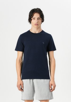 Navy blue cotton t-shirt with a round neck and short sleeves, featuring a small logo on the chest. Model wears striped shorts.