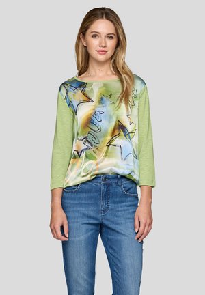 Green three-quarter sleeve top with a colorful star pattern and abstract designs; paired with blue denim jeans. Smooth texture, relaxed fit.