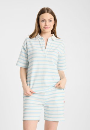 Młoda kobieta w jasnoniebiesko-białej koszulce polo w paski z krótkim rękawem i pasujących szortach, stojąca na jednolitym białym tle.