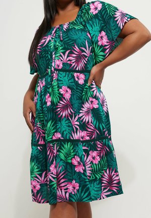 Kvinde iført grøn kjole med pink og grøn tropisk blomsterprint, korte ærmer og rynket halsudskæring, stående med hænderne på hofterne.