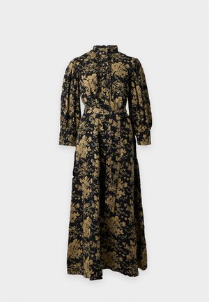 Femme aux cheveux bouclés mi-longs portant une longue robe noire à motif floral doré et des chaussures plates beige clair, debout sur fond blanc.