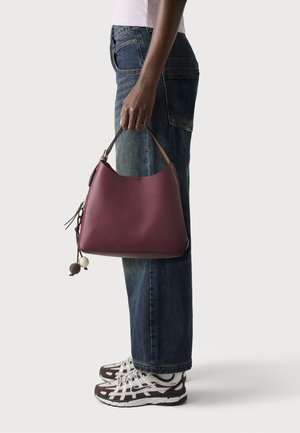 Person holder en lilla lædertaske med en kort brun rem, iført blå jeans, hvid top og sorte og hvide sneakers.