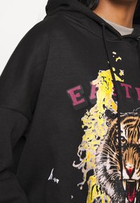 Svart huvtröja med framsidan tryckt en rytande tiger omgiven av gula stänk och delvis synlig stor röd text.