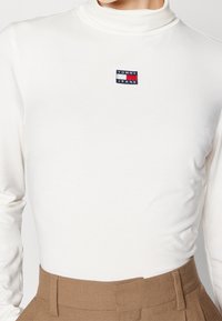 White turtleneck top made of soft fabric, features a small blue, red, and black embroidered logo patch on the chest. Fitted design.