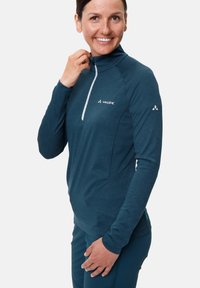 Pull-over teal avec une fermeture éclair quart et des manches longues. Tissu léger avec une texture lisse. Présente un logo sur la poitrine et l'épaule.