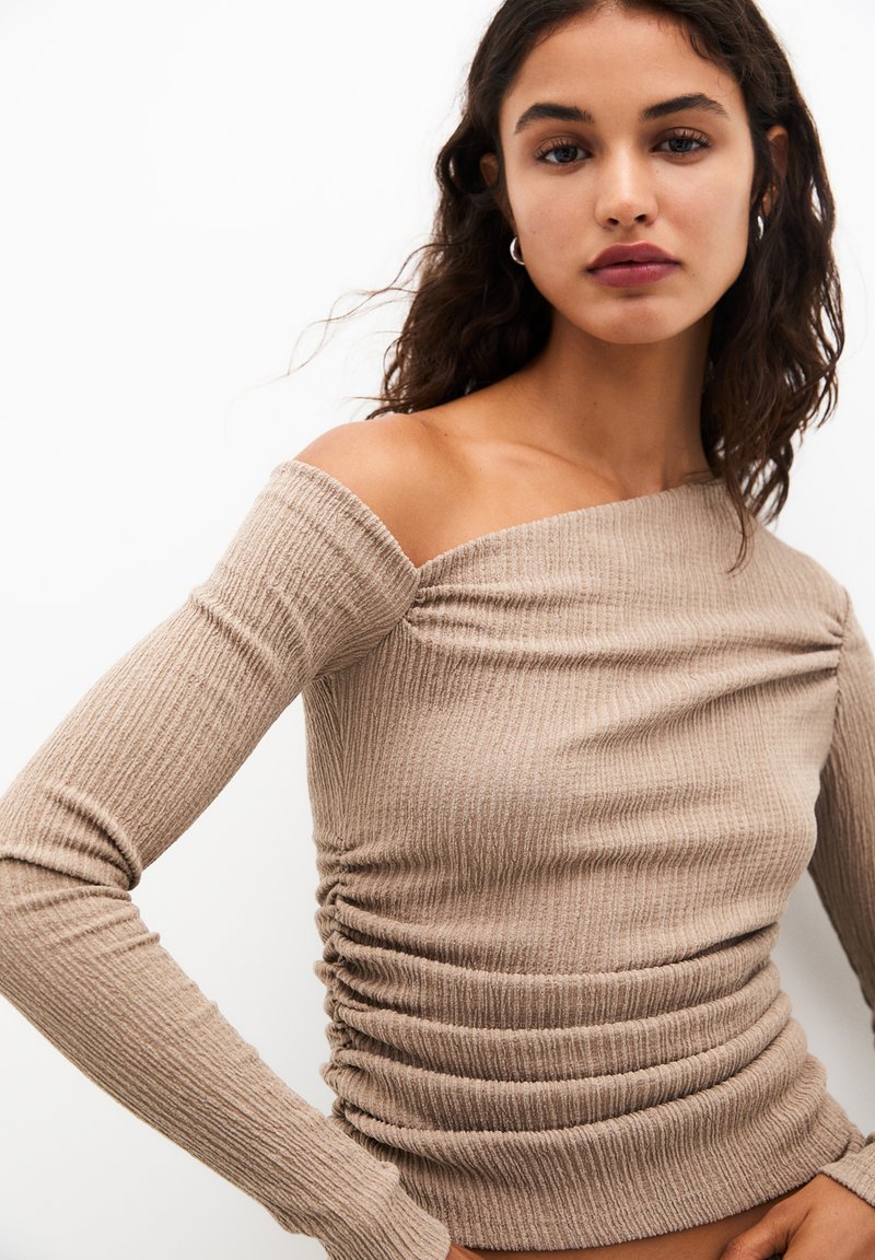 Top long bleu beige à manches longues avec une épaule découverte, présentant des détails froncés sur le côté pour une texture supplémentaire.