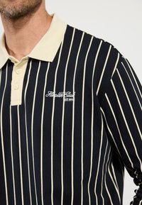 Marineblauwe polo met lange mouwen en verticale crèmekleurige strepen, voorzien van een crèmekleurige kraag en geborduurd "Health Club" logo op de borst.