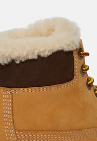 Stivale in pelle marrone chiaro con cavigliera in suede marrone, collare in shearling crema, lacci gialli e rossi, e dettagli in cucitura bianca.
