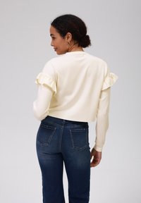 Top corto color crema con maniche a volant abbinato a jeans a vita alta blu scuro. Texture liscia e design aderente.
