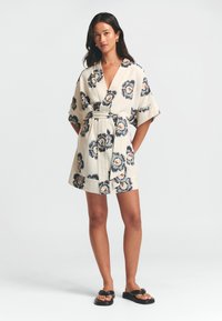 Krátké rukávy, zavinovací šaty v světle béžové látce s květinovým vzorem v tmavě modré a oranžové barvě. Obsahují ladící opasek. Ploché černé sandály.