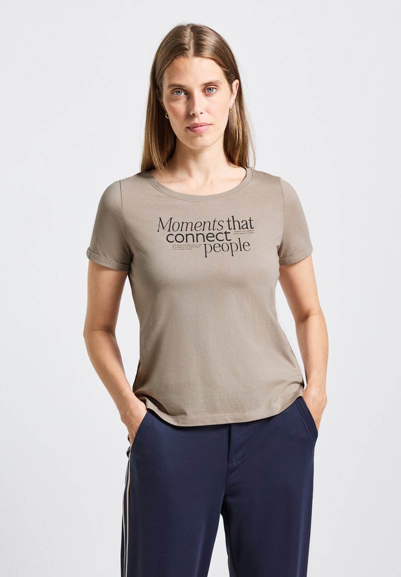 Camiseta de manga corta en color verde oliva con un cuello redondo y texto en negro: "Momentos que conectan a las personas." Tejido suave y liso.