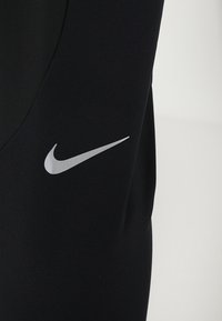 Černé sportovní oblečení Nike s hladkou texturou, přiléhavým střihem a výrazným stříbrným logem na levé straně.