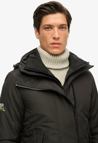 Zwarte zip-up jas van een gestructureerde stof, met een capuchon, zijzakken en een logodetail. Lichtgewicht met een gladde afwerking.