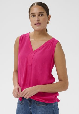 Femme aux cheveux foncés tirés en arrière, portant des boucles d'oreilles créoles dorées, un chemisier sans manches rose vif à col en V, et un jean bleu, debout devant un fond neutre.