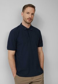 Navyblauwe polo met korte mouwen, met een gestructureerd ruitmotief, een sluiting met drie knopen en een kraag. Draagt een combinatie met kaki broek.
