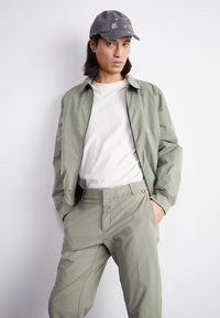 Jachetă bomber verde deschis cu fermoar, tricou alb cu guler rotund și pantaloni asortați. Căciulă gri cu detalii grafice subtile.