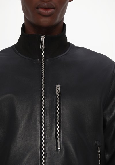 Μαύρο δερμάτινο μπόμμερ μπουφάν με ψηλό γιακά, που διαθέτει ασημένιες φερμουάρ και μπροστινή τσέπη. Λείος υφασμάτινος χαρακτήρας και εφαρμοστή σχεδίαση.