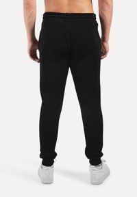 Muž stojící naboso, otočený zády, v černých teplácích a bílých teniskách na jednobarevném bílém pozadí.