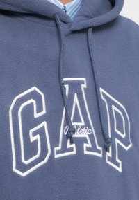 Pogled od blizu modre jopa z bombažno podlogo, na kateri je napis »GAP Athletic« v beli barvi in vrvice za zategovanje, čez ovratnik modre srajce s črtami.