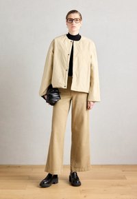 Beige, cropped jacket with snap closures over a black turtleneck; wide-legged tan trousers; black pleated handbag; chunky black shoes.
