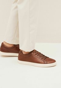 Bruine leren sneakers hebben een gladde textuur, een ronde neus en een witte rubberen zool. Veters in bijpassend bruin bevestigen het ontwerp.