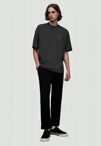 Young man with shoulder-length hair wearing a loose dark gray T-shirt, black trousers, and black sneakers with white soles and leopard print.