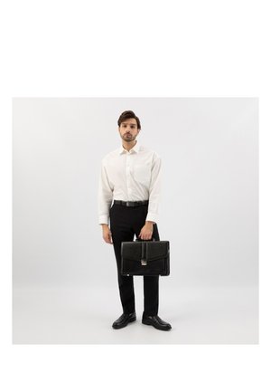 Man in wit shirt en zwarte broek die een zwarte leren aktetas vasthoudt, staande tegen een effen lichtgrijze achtergrond.