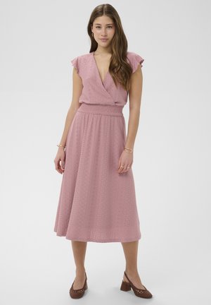 Mladá žena stojaca v ružových midi šatách s dierkovaným vzorom, s rozstrapkanými rukávmi a hnedými topánkami na hrubom opätku na jednoduchom pozadí.