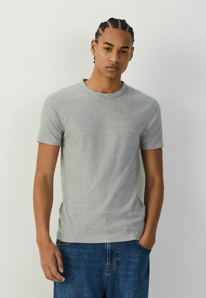 Jonge man met gevlochten haar die een eenvoudige grijze T-shirt met korte mouwen en een blauwe spijkerbroek draagt, staand tegen een effen lichte achtergrond.