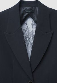 Schwarzer Blazer mit breiten Revers und sichtbarem Innenfutter, das ein zartes florales Muster aus schwarzer Spitze aufweist.