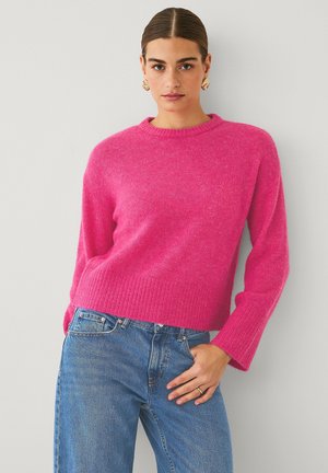 Fuchsia roze gebreide trui met ribbelrand en brede mouwen, gedragen met hoge spijkerjeans in het blauw. Soepele textuur, casual pasvorm.