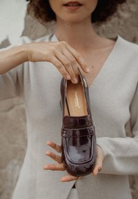 Mocasín de charol burdeos con punta cuadrada, detalle de correa en la parte frontal y un acabado liso y brillante, sujetado por una persona con un suéter claro.