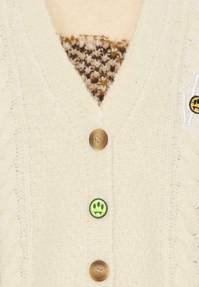 Beżowy sweter na drutach z wzorem warkocza, z trzema brązowymi guzikami i zieloną naszywką z uśmiechniętą buźką na przodzie. Brązowa i kremowa wzorzysta warstwa wewnętrzna.