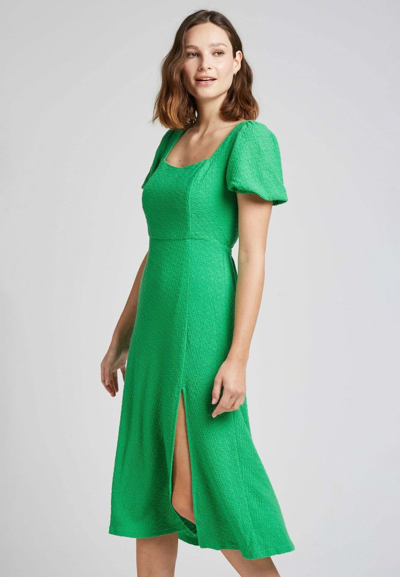 Robe midi texturée verte avec des manches courtes à volants, décolleté carré et fente latérale jusqu'à la cuisse. Tissu lisse et design ajusté.