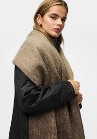 Bred, varm sjal i en gradient af brune nuancer, lavet af blød uld, båret over en sort, quiltet jakke. Har et draperet design og tekstureret stof.