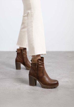 Botas de cuero marrón hasta el tobillo con un alto tacón cuadrado, cremallera lateral y ribete de piel texturizada en la parte superior. Pantalones blancos recortados con dobladillo deshilachado.