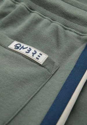 Pantaloni jogger in misto cotone verde con vita a trama, caratterizzati da un'etichetta bianca con testo blu e strisce laterali in blu navy e bianco.