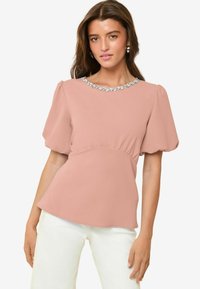 Blusa color rosa con maniche a sbuffo, vita arricciata e dettagli di gioielli sul collo. Il tessuto morbido conferisce una texture delicata.