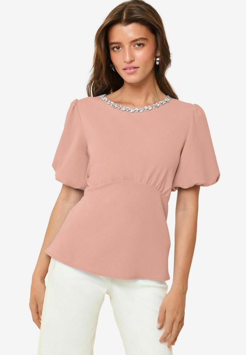 Blusa color rosa con maniche a sbuffo, vita arricciata e dettagli di gioielli sul collo. Il tessuto morbido conferisce una texture delicata.