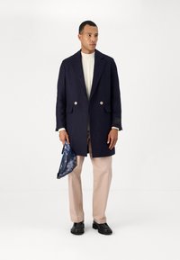 Palton marin cu două rânduri de nasturi mari, pulover crem cu model de tricotaj, pantaloni bej și o geantă de mână albastră cu model. Botine negre.