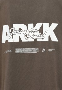 Brązowa bawełniana koszulka z wyraźnym białym logo "ARKK" oraz napisem. Napis stylizowany jest w mieszance czcionek blokowych i kursywy.