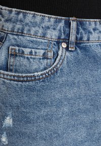 Un jean en denim bleu clair présente un détail déchiré, des coutures visibles et un design classique à cinq poches avec une fermeture à bouton et des passants de ceinture.