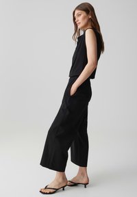 Young woman in sleeveless black top, wide-leg black pants, posing in profile with hands in pockets against plain background.
