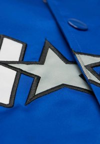 Blaue Jacke mit großen weißen und grauen Stern-Design. Besitzt schwarze Nahtdetails und einen Knopfverschluss. Glatte Stoffoberfläche.
