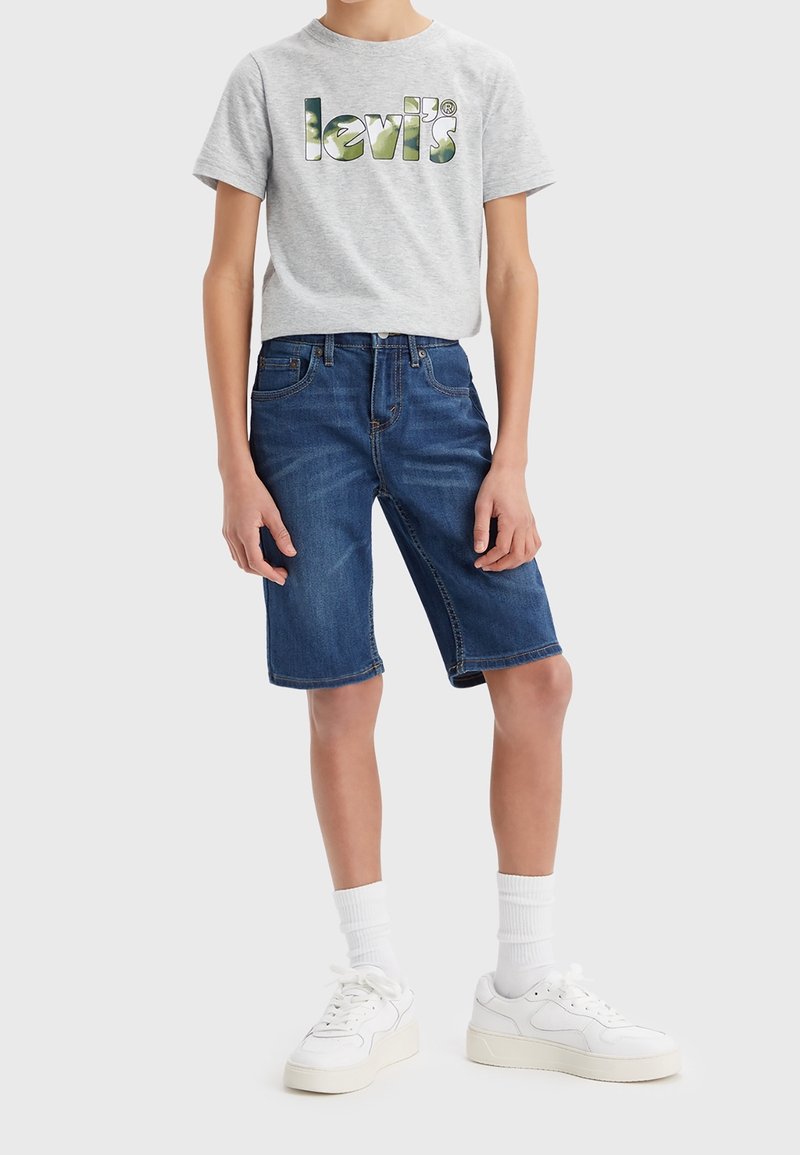 Camiseta gris con el logo de "Levi's" en verde, combinada con pantalones cortos de mezclilla azul. Zapatillas blancas y calcetines blancos de canalé completan el atuendo.